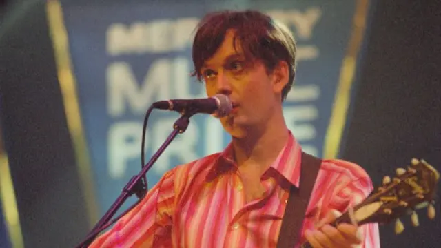 Pulp singer Jarvis Cocker performing on stage at the 1996 Mercury Prize awards, wearing a pink stripy shirt and holding a guitar