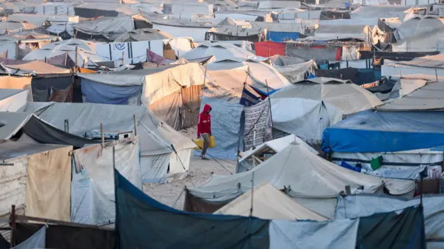 A Palestinian carrying containers walks among tents in Khan Younis