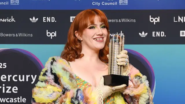 Irish singer CMAT arrives at this year's Mercury Prize awards in Newcastle. She's in a multi-coloured tulle frock smiling as she holds up her shortlisted album Mercury Prize award