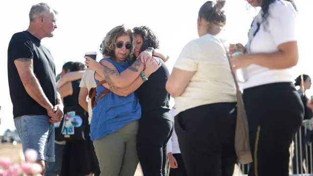 Two women in the centre of the image embrace. A man watches with clasped hands on the left, while two other women are watching from the right