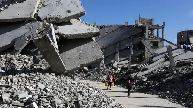 A man carrying two yellow plastic jerry cans walks down a road, a young boy ahead of him. Around them are the debris and remains of several collapsed buildings