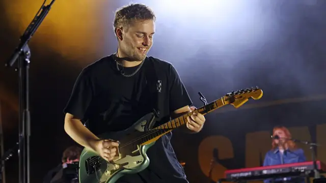 Artist Sam Fender smiling and playing the guitar on stage.