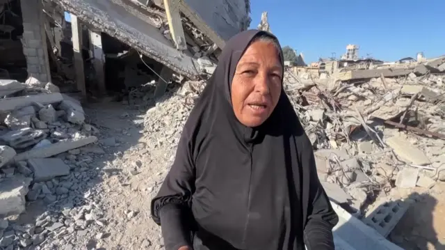 Hayam Meqdad in a black abaya speaks to the camera, rubbles and debris of damages homes all around her
