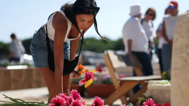 A woman in denim shorts and white vest laying flowers down on a grave
