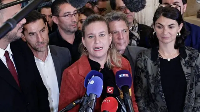 Mathilde Panot in a red jacket speaks to reporters in front of microphones