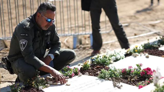 A man in police or military uniform and sunglasses kneels next to a grave and rests the fingertips of his right hand on it