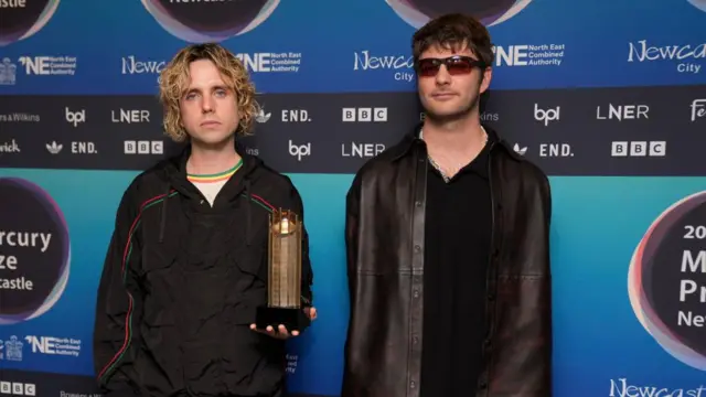 Two men wearing black clothing, one holding a cylinder shaped award