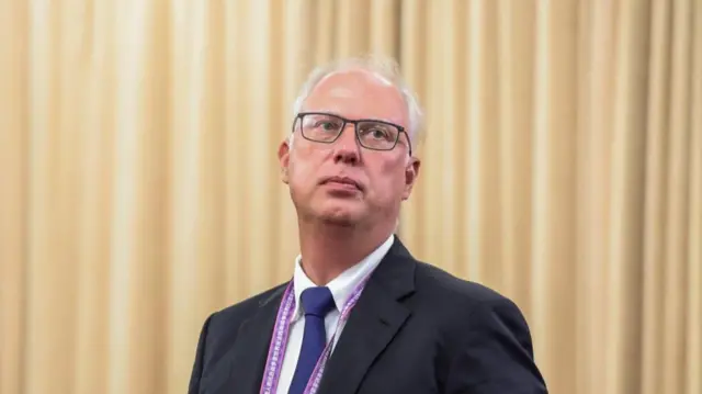 A man in a black suit in front of a yellow curtain