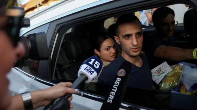 A man sits in a car and looks out the window at the microphones of reporters being pointed towards him