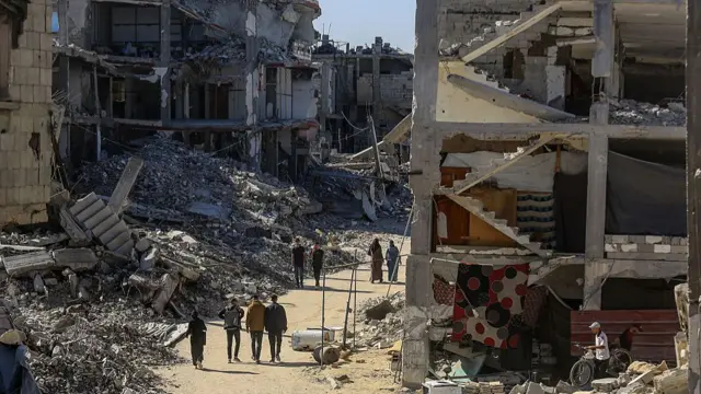 People walk a path through huge piles of rubble, in the foregound of the picture a shell of a house with the internal hallway exposed is visible