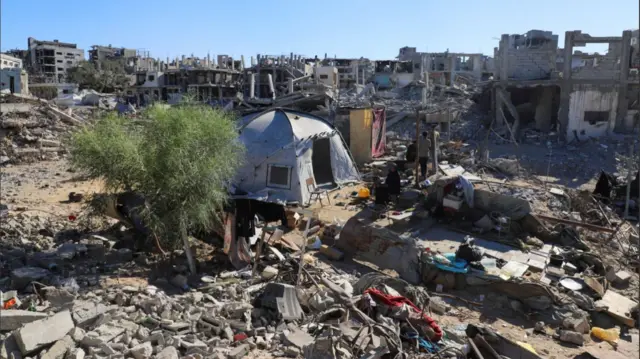 Hayam Meqdad sits on a plastic chair outside a grey tent pitched on top of the ruins of her destroyed home in Gaza City. In the background, several damaged and destroyed buildings