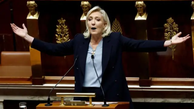 A woman with blond hair gestures with her hands held out to her sides