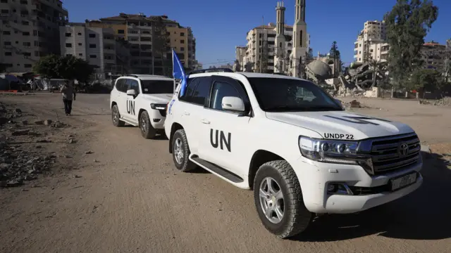 Two white UN Development Programme vehicles pictured outdoors in Gaza. In the background are buildings, some intact and some damaged