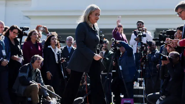 Karoline Leavitt wears a blazer and walks away from a microphone with cameras in the background