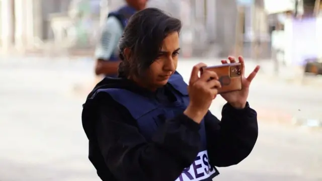Woman in navy press vest holds a phone up to take a photo of something in front of her