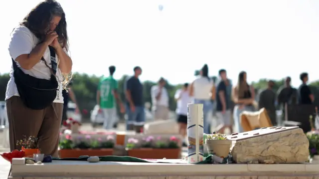 A woman holding her clasped hands to her mouth as she looks down at a grave. She is wearing a white tshirt and brown trousers.