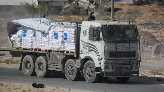 Truck loaded with white aid sacks that have blue World Food Programme labels. It is travelling on a road, with a person in a black hooded jumper and blue surgical mask sitting with the aid.
