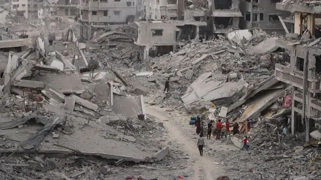A group of people stand on a dusty path that goes through huge piles of debris and ruined buildings
