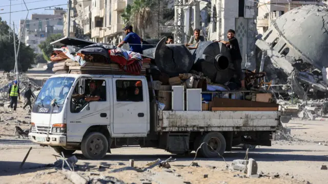 Palestinians ride on a truck loaded with belongings in Gaza City, with destroyed buildings in the background