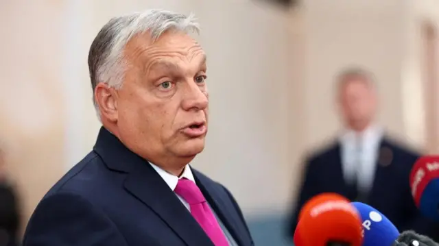 Viktor Orban in a blue suit and pink tie stands at a microphone
