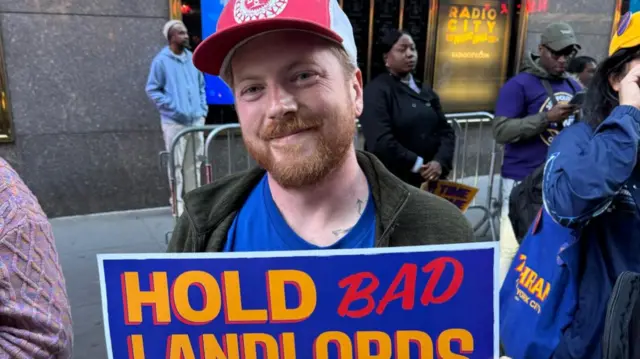 Miles Ashton wears a white and red hat and a blue shirt. He stands in front of Rockefeller Center holding a sign that says "hold bad landlords accountable"