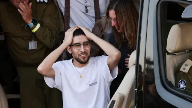 Released hostage Evyatar David holds hand on head and smiles upon arrival at Rabin Medical Center. He's standing in the entranceway of a helicopter with people standing behind him