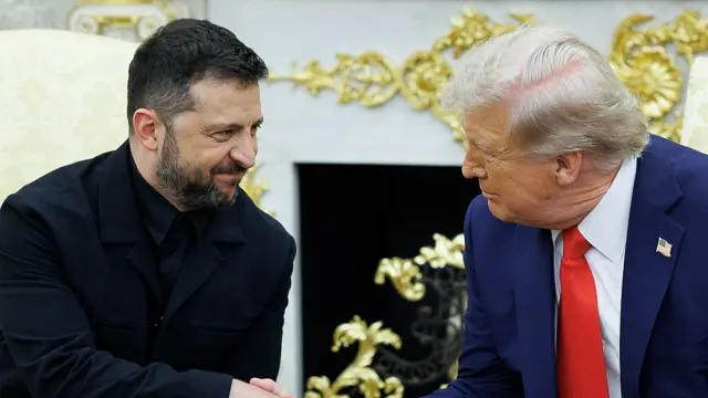 Volodymyr Zelensky (L) smiles with his mouth closed as he shakes hands with Donald Trump (R). They're sitting inside the Oval Office
