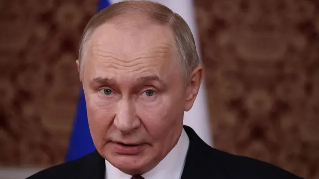 close-up of Vladimir Putin in a black suit and white shirt, looking slightly to the left, behind him a Russian flag with the blue and white parts visible