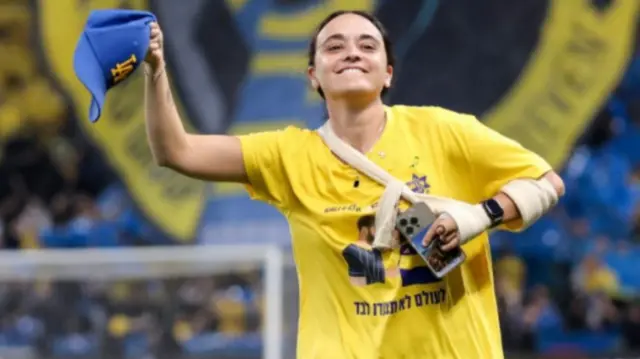 Emily Damari waves a baseball cap on the pitch of Maccabi Tel Aviv - gesturing towards the crowd of her beloved football team
