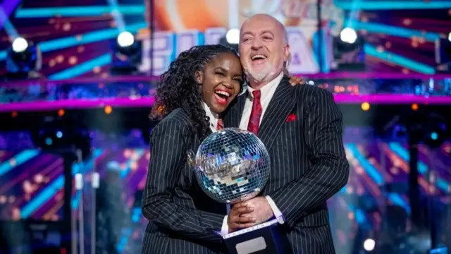 alt="Oti Mabuse and Bill Bailey in dark, pinstripe suits and red ties hold the Glitterball Trophy after winning series 18 of Strictly Come Dancing."
