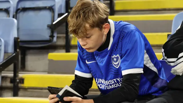 A young Portsmouth fan checks his phone while sat in the stands at the King Power Stadium