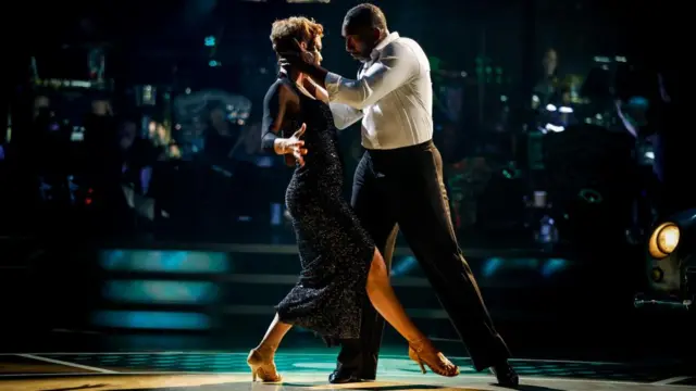 A woman in a black dress with short brown hair dances with a man in semi-formal wear.