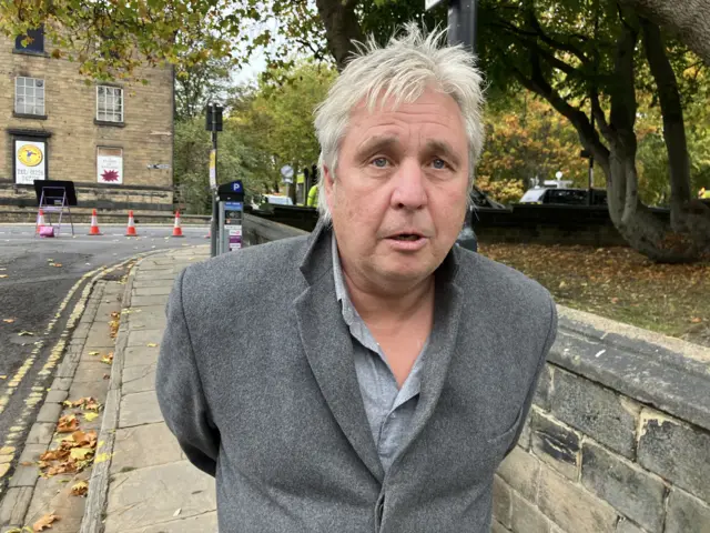 A man with white hair stood on a street wearing a blazer and a shirt.