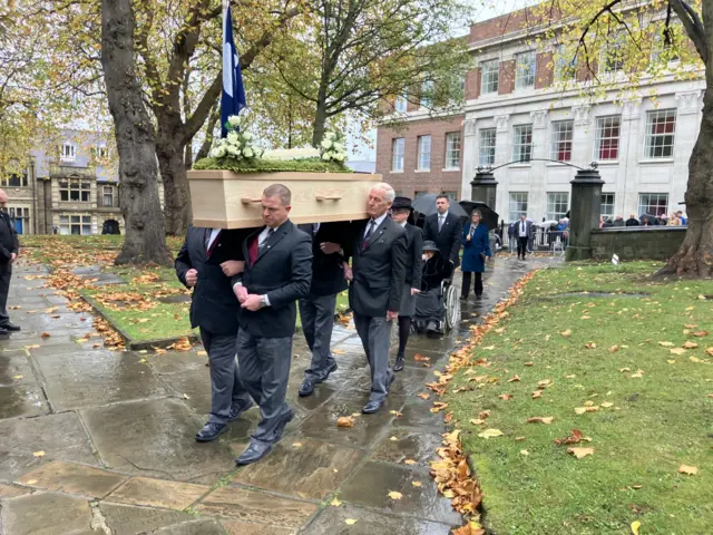 A coffin carried by pallbearers with mourners following behind