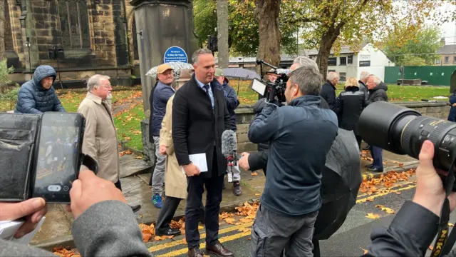 A man being interviewed outside a church with people walking past