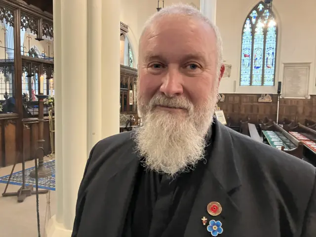 A reverend with a white beard smiling inside a church`