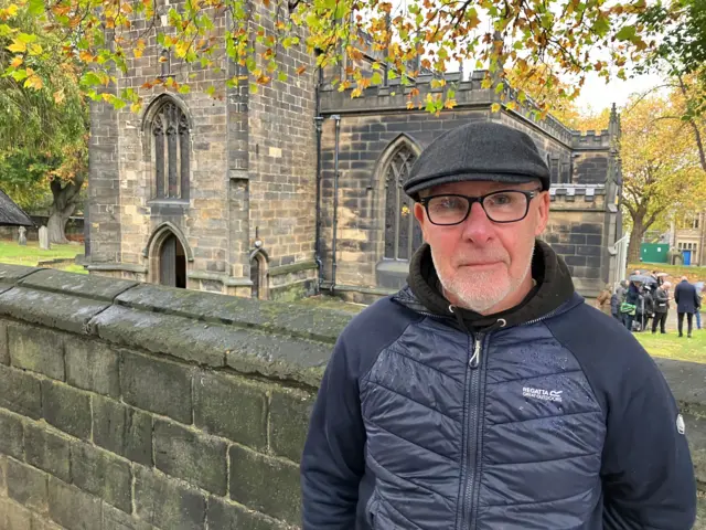 A man wearing glasses and a flat cap standing in front of a church