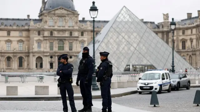 Three police officers stand outside the Louvre's famous glass pyramid entrance