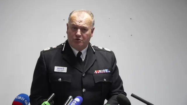 Sir Stephen Watson delivers announcement in full black uniform with white wall behind him
