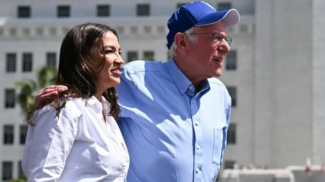 Ocasio-Cortes and Sanders stand together. Sanders is in a blue shirt and hat with his arm around Ocasio-Cortes, who wears a white shirt