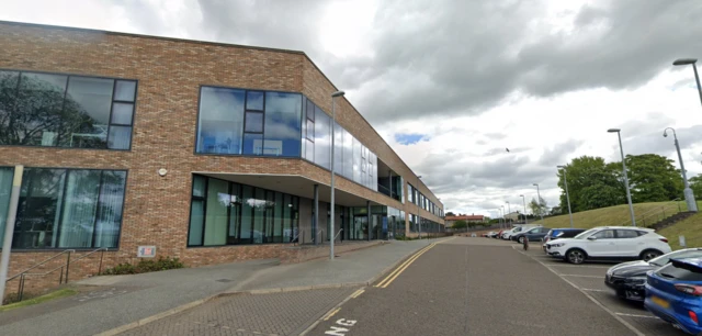 Forth Valley College campus in Alloa, a modern two-story brick building with large glass windows and a covered entrance. The building is adjacent to a road lined with parked cars on the right side. A grassy slope with steps and trees is visible in the background under a cloudy sky."