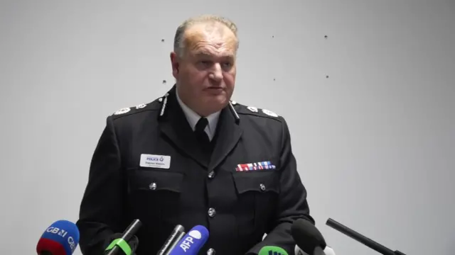 Chief Constable Sir Stephen Watson addresses press in full uniform, several mics in front of him and a white wall behind him