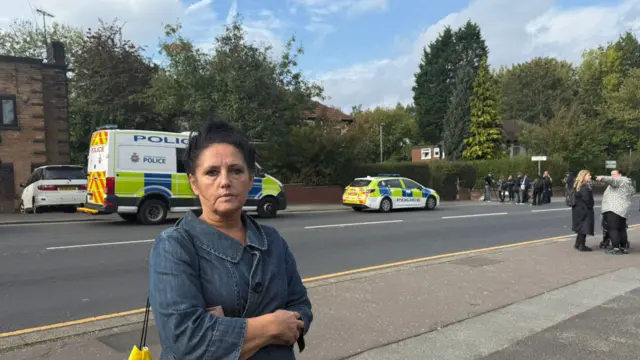 Claire has dark brown hair tied in a bun on top of her head, and is wearing a denim jacket. There is a police van and car parked on the road behind her.