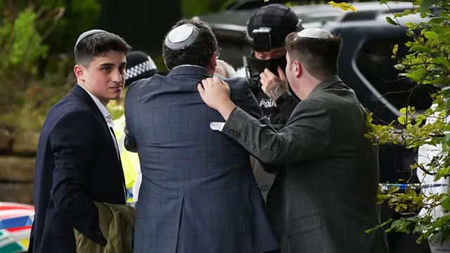 Members of the Jewish community comfort each other, while a heavily armed police officer can be seen in the background