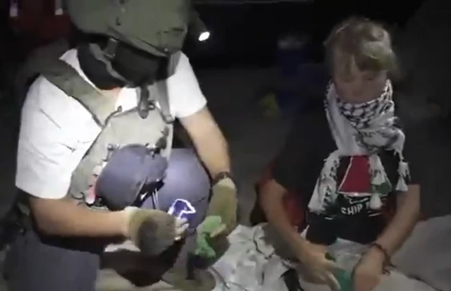 Screengrab showing a man wearing a helmet, bulletproof vest and face covering squatting next to a seated Greta Thunberg. She is wearing a Keffiyeh scarf.