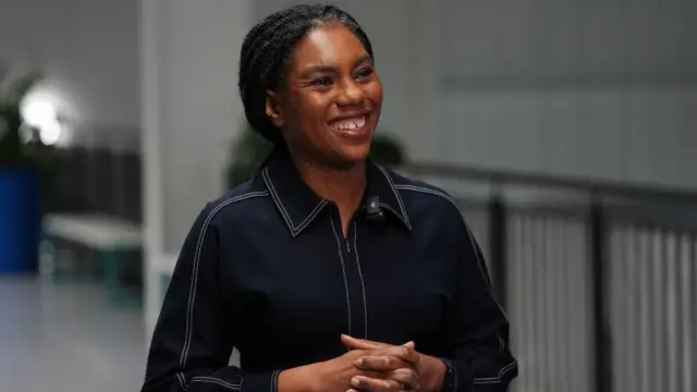 Kemi Badenoch smiles and wears a black dress shirt.