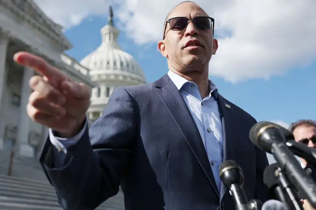 House Minority Leader Hakeem Jeffries speaks on the steps of the US Capitol whose dome is behind him. He is wearing a navy blue suit over a light blue shirt and black sunglasses.