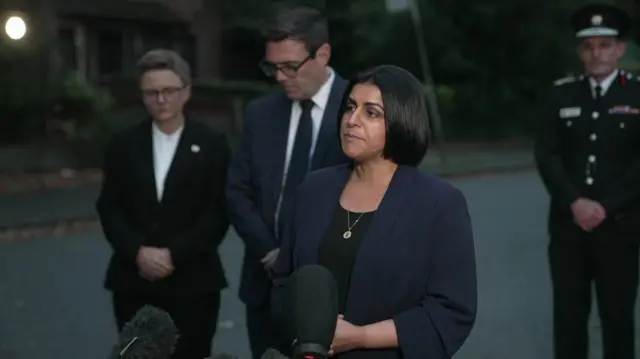 Home Secretary Shabana Mahmood gives statement wearing black blazer and top, Andy Burnham behind her in black suit and head down