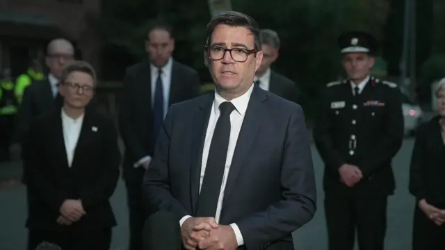 Andy Burnham speaking to the media, there are police officers behind him.