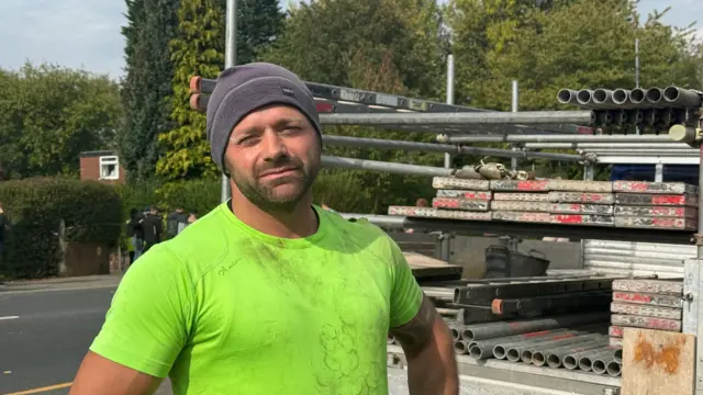 Mark Farlow, a scaffolder from Ashton. He is wearing a neon green top and a grey beanie. To his right there is a car with scaffolding equipment on it.